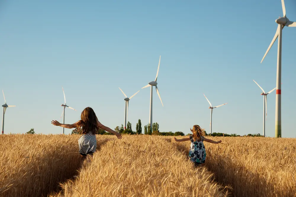 Girls run through a field towards wind turbines.